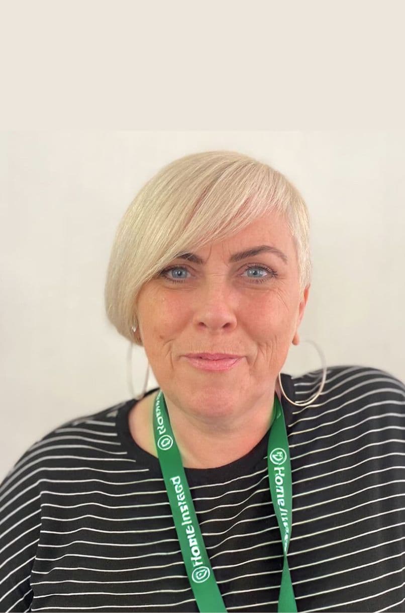 Woman with short blonde hair, striped shirt, hoop earrings, and a green "Home Instead" lanyard, smiling. - Home Instead