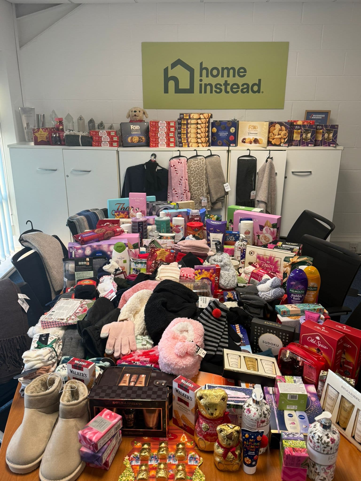 A table filled with assorted gifts and treats under a "Home Instead" sign in an office setting. - Home Instead