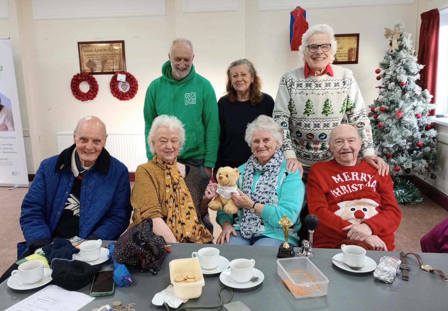 Seven smiling pensioners, some in festive jumpers, gather around a table by a decorated Christmas tree. - Home Instead