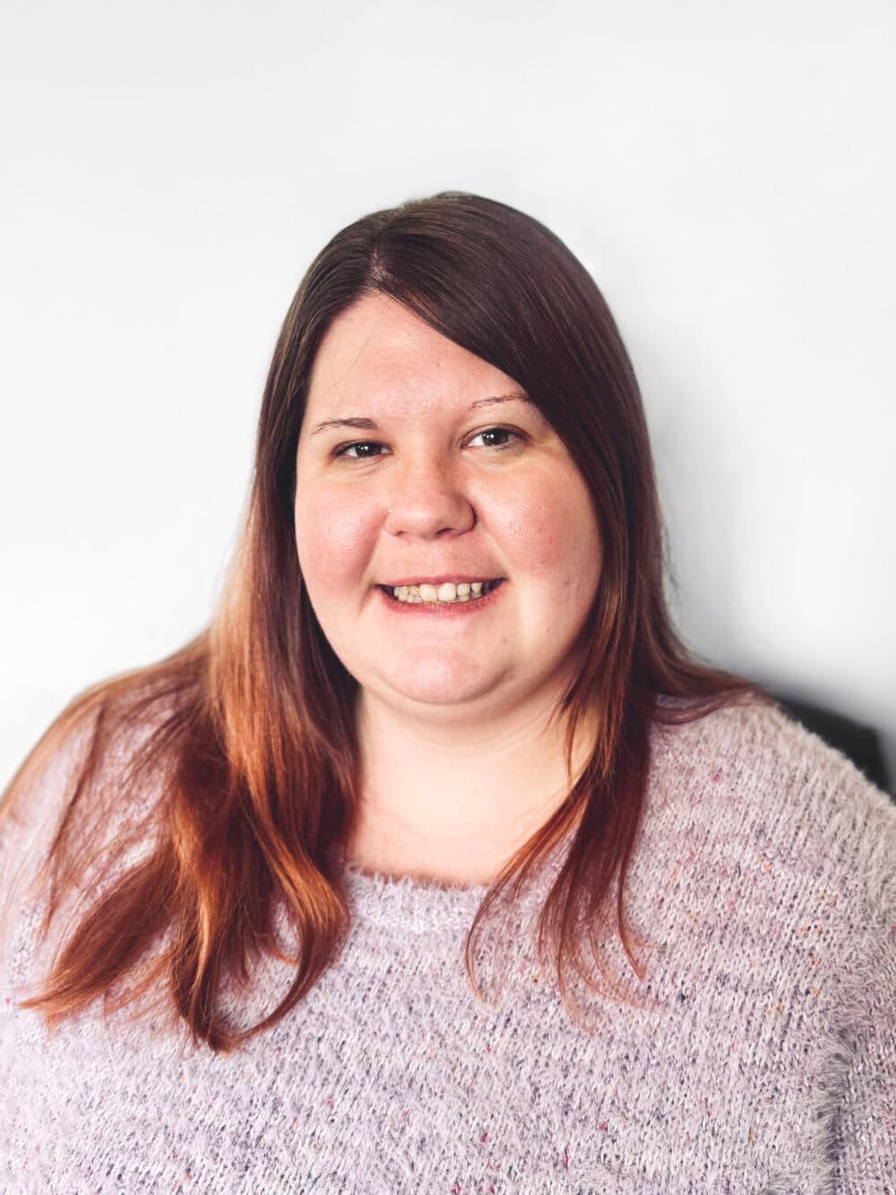Smiling woman with long brown hair wearing a light purple sweater against a plain white background. - Home Instead