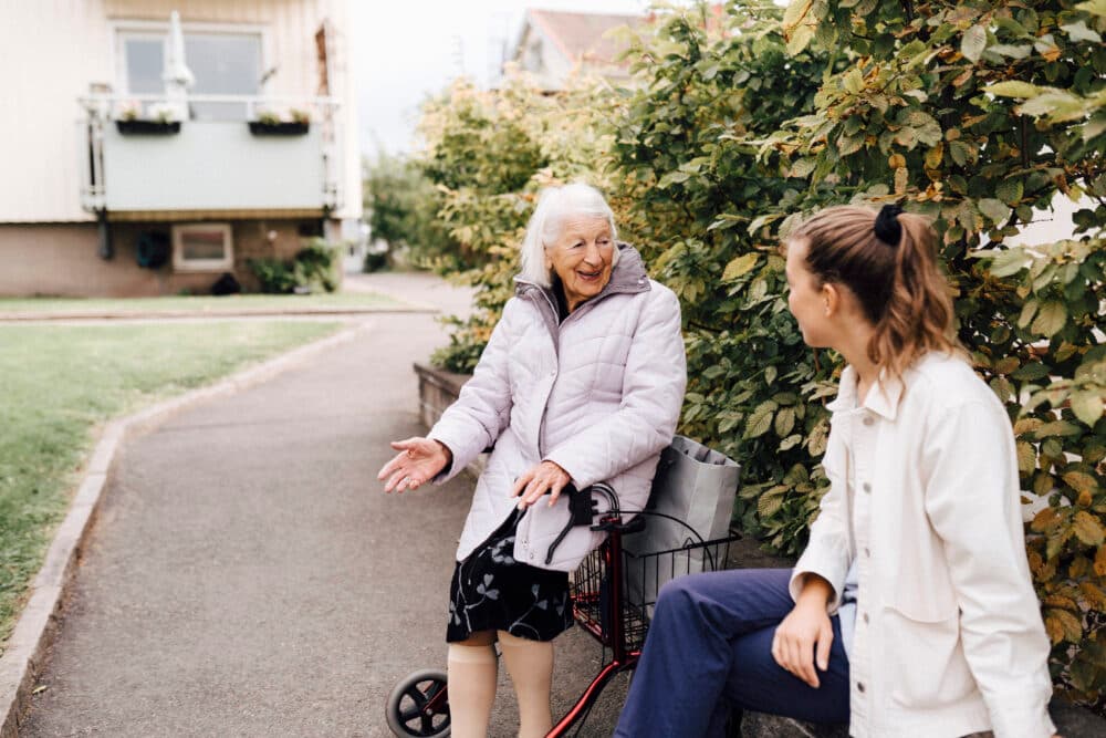 An elderly woman sits on a walker, smiling and talking with a young woman outdoors near green bushes. - Home Instead