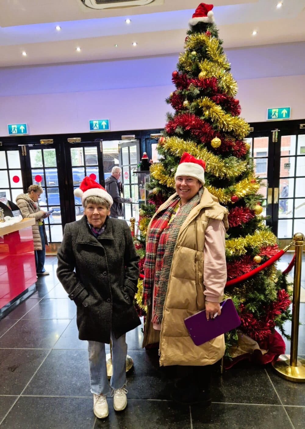 Two women in Santa hats pose by a decorated Christmas tree in a festive indoor setting. - Home Instead