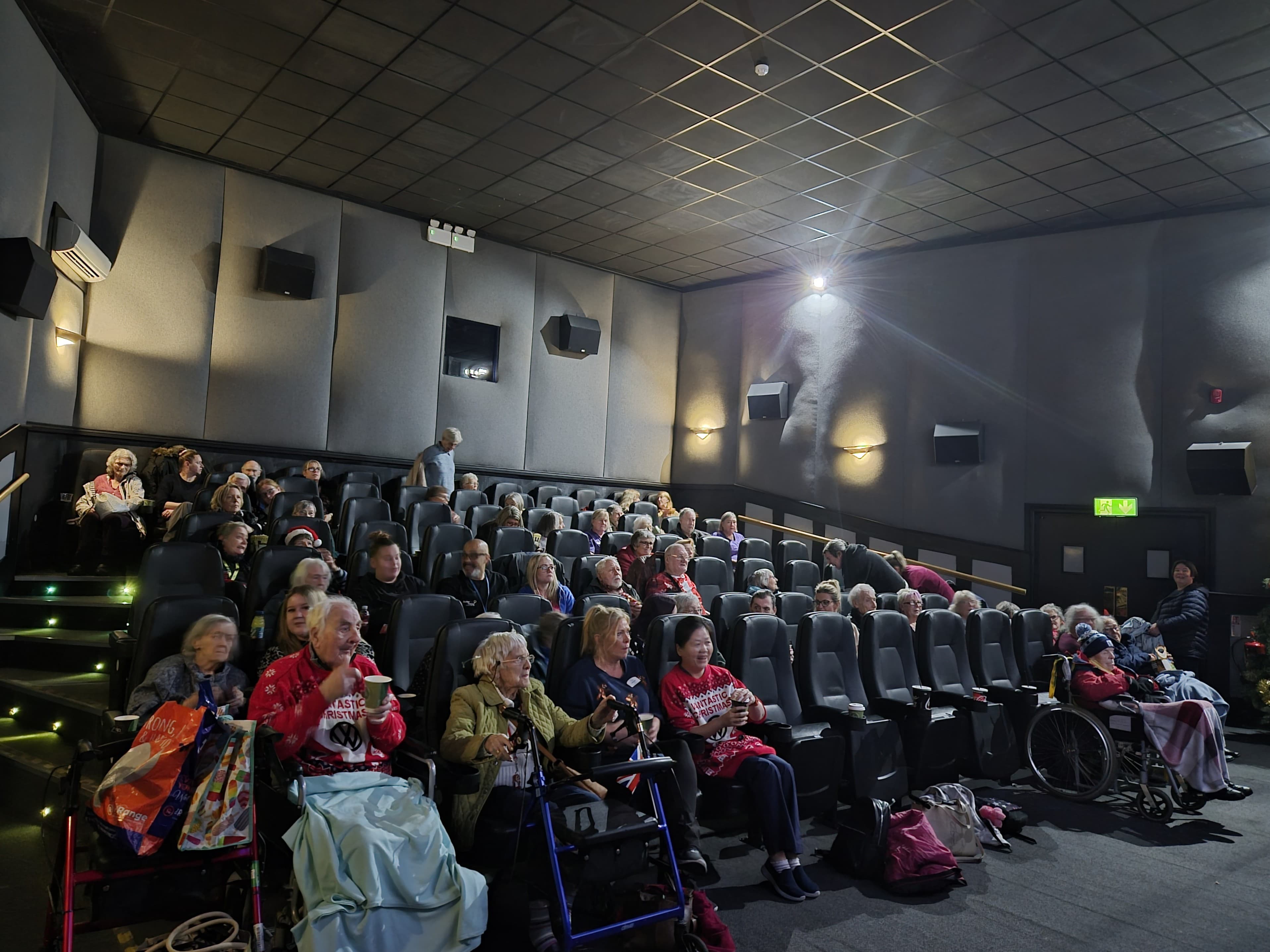 Elderly people sitting in a dark cinema, some with walking frames and wheelchairs, waiting for a film to start. - Home Instead