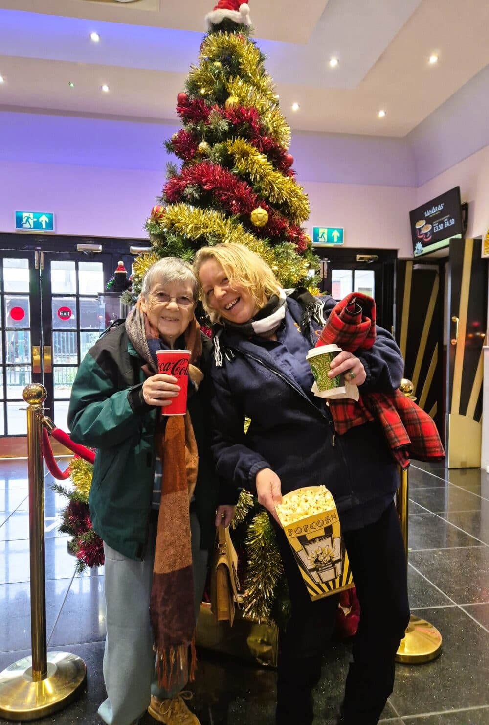Two women smiling with popcorn and drinks in front of a decorated Christmas tree indoors. - Home Instead
