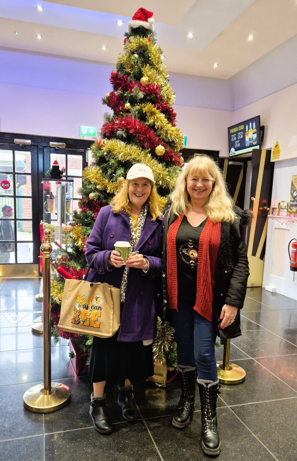 Two women smiling in front of a decorated Christmas tree indoors, holding a drink and a bag. - Home Instead