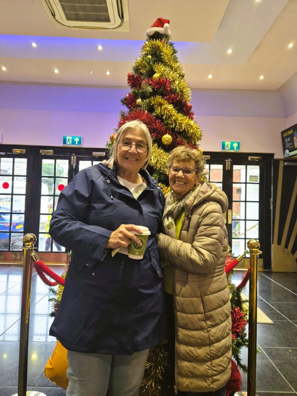 Two smiling women stand in front of a decorated Christmas tree indoors, one holding a takeaway coffee cup. - Home Instead