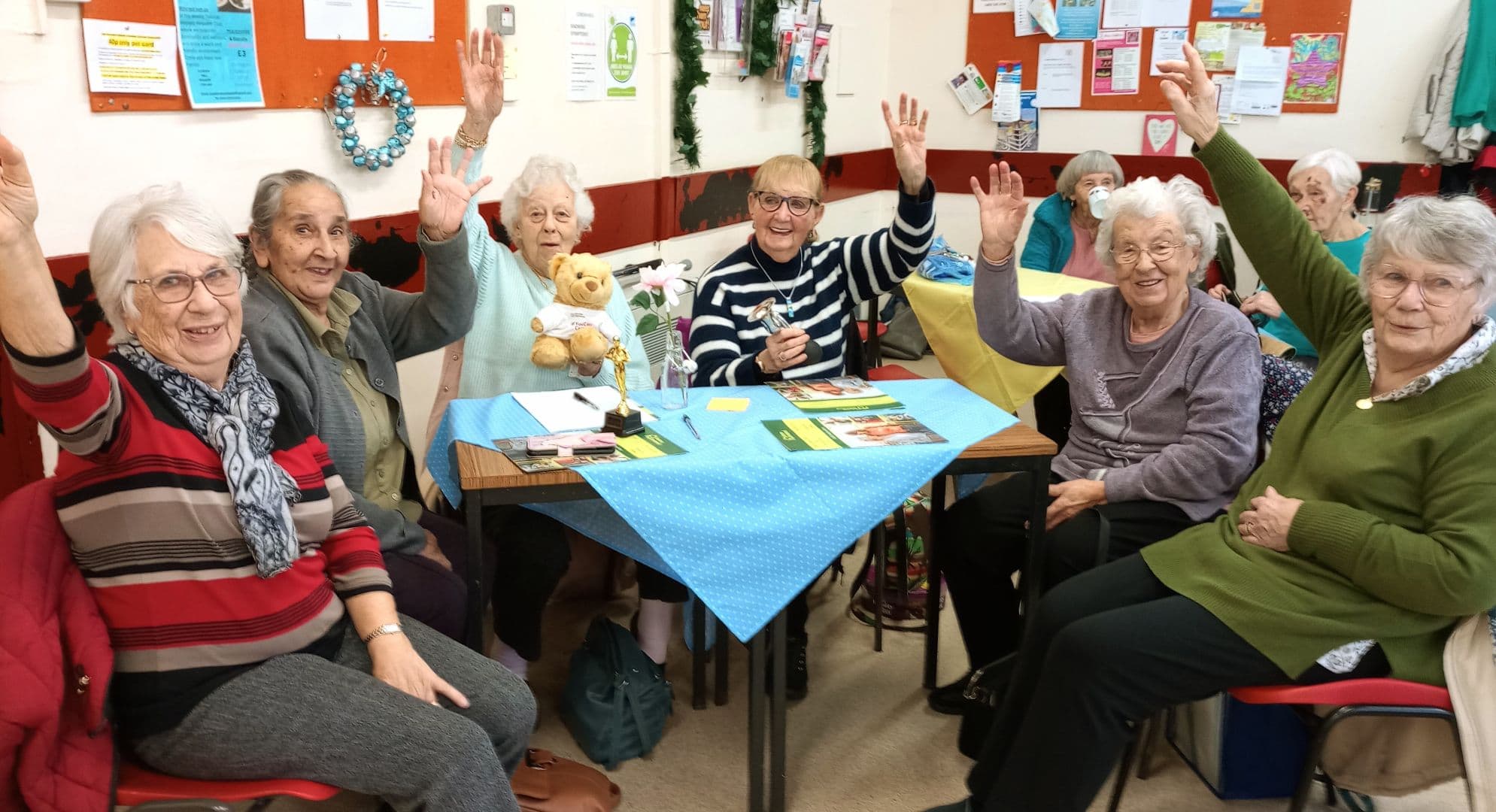 A group of smiling seniors at a community center raise their hands around a table with crafts and snacks. - Home Instead
