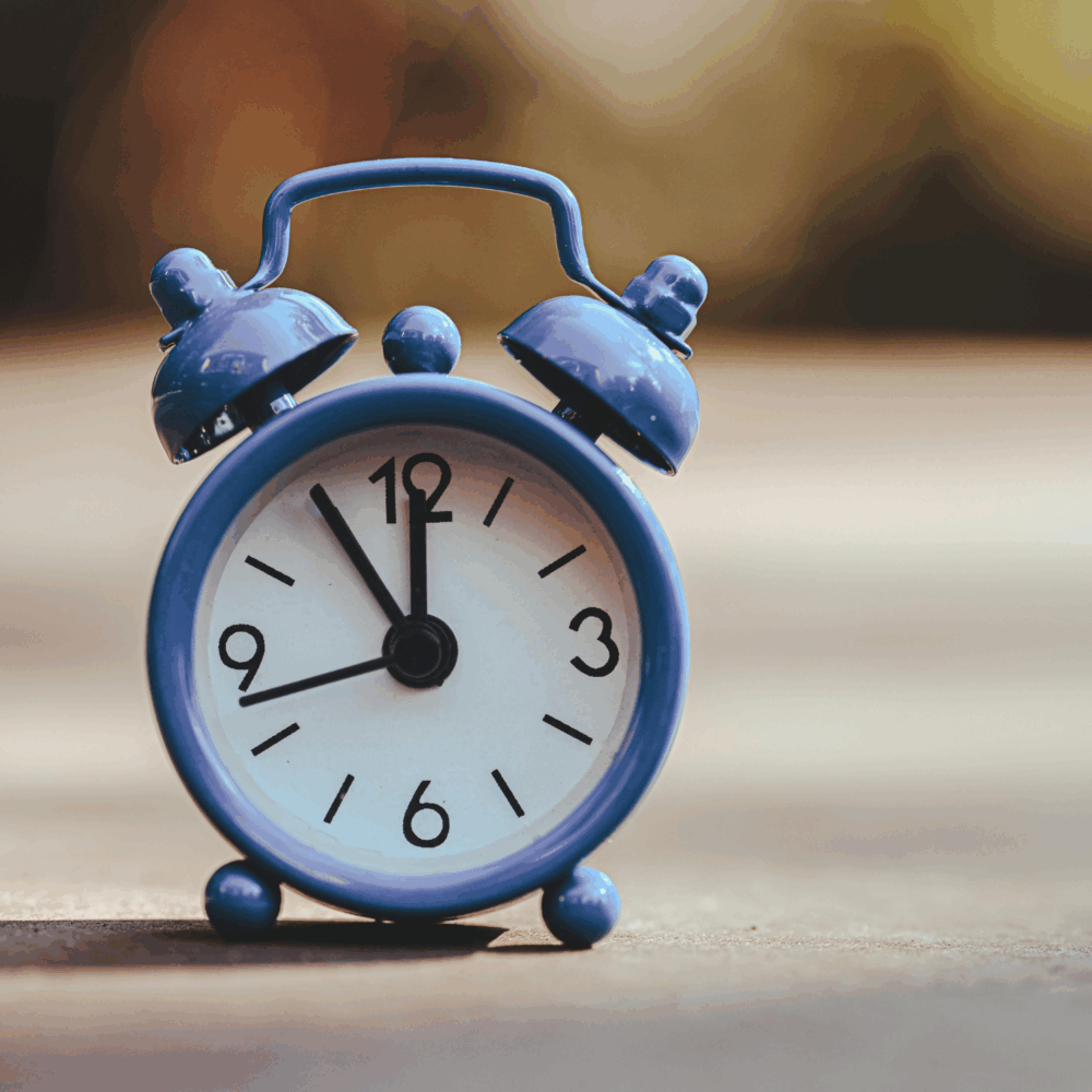 A blue analogue alarm clock showing 7:00, placed on a smooth surface with a blurred background. - Home Instead
