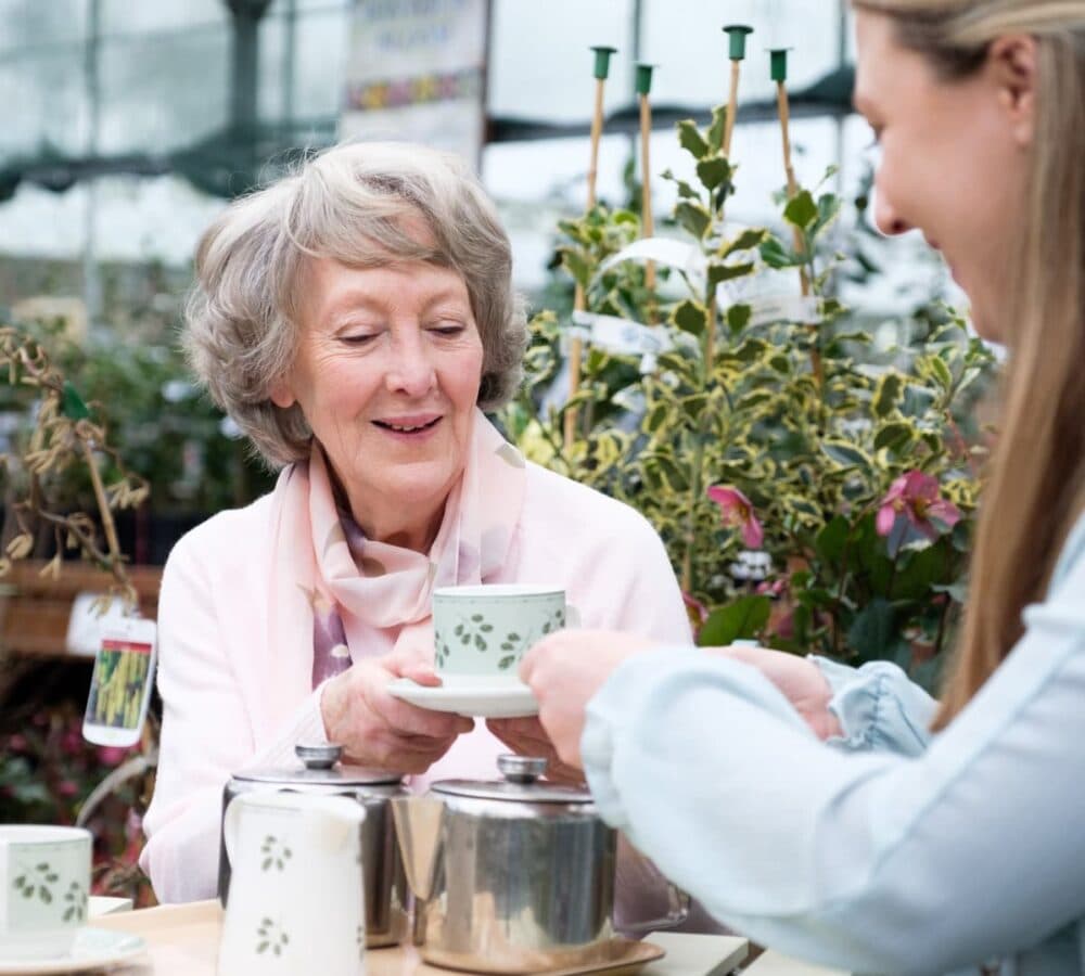 An older female adult with grey hair having a cup of tea with her female carer outdoors