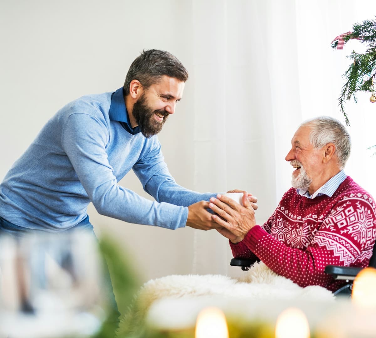 An older male adult with grey hair wearing red Christmas sweater and receiving a gift while sitting on a wheelchair with a man with beard and wearing blue both happy and smiling