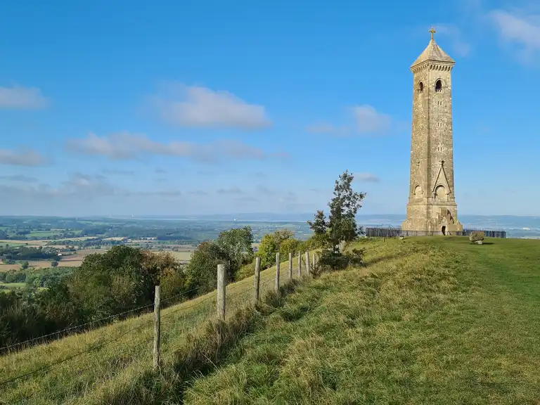 Tall stone tower on a grassy hill with fence, trees, and countryside under a blue sky with clouds. - Home Instead