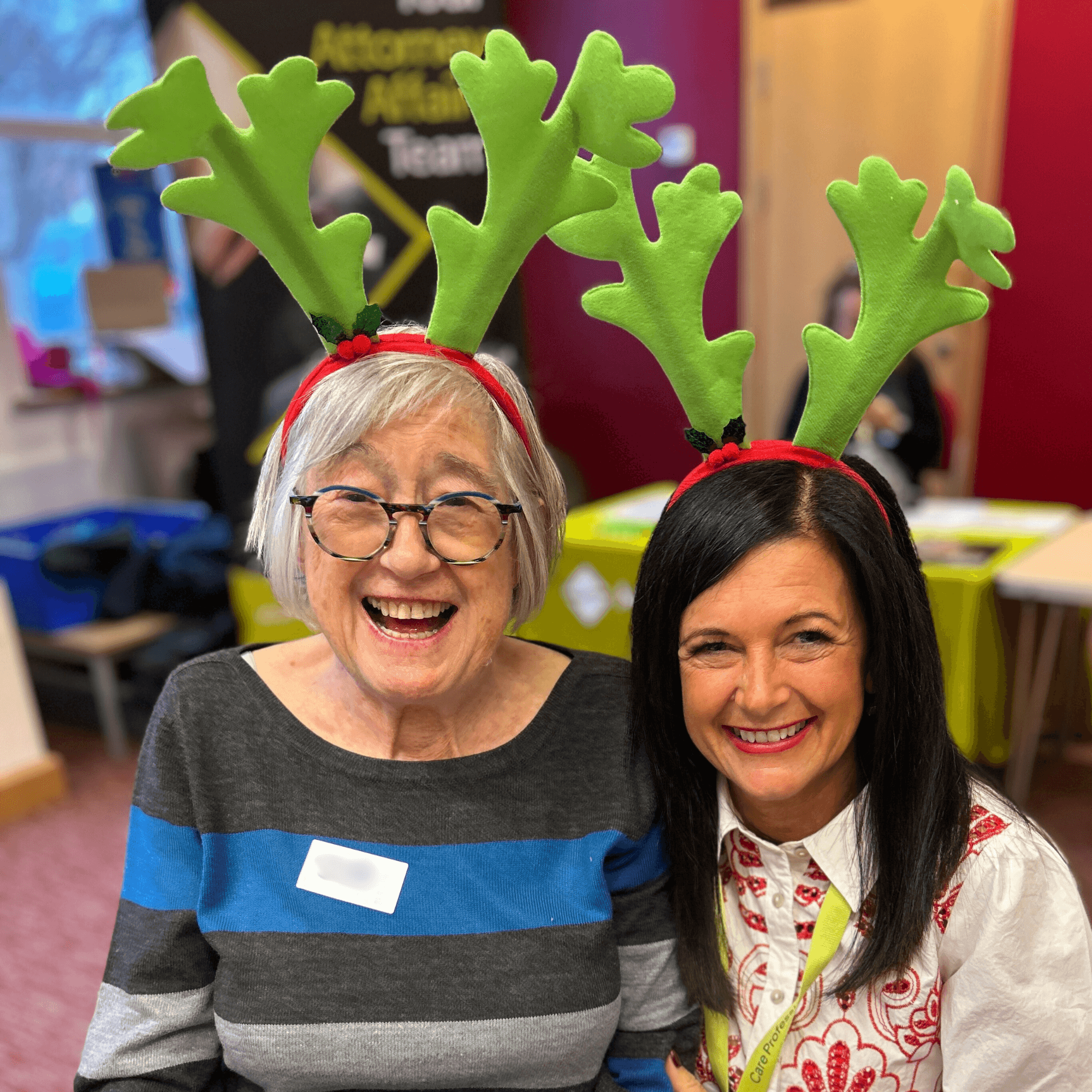 Two women smiling at a table, both wearing green reindeer antler headbands at a festive indoor event.