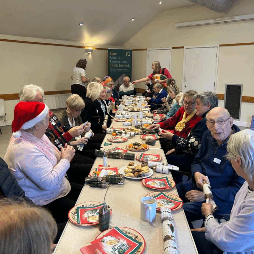 A group of elderly people enjoy a festive meal together at a long table, wearing Christmas hats and jumpers. - Home Instead