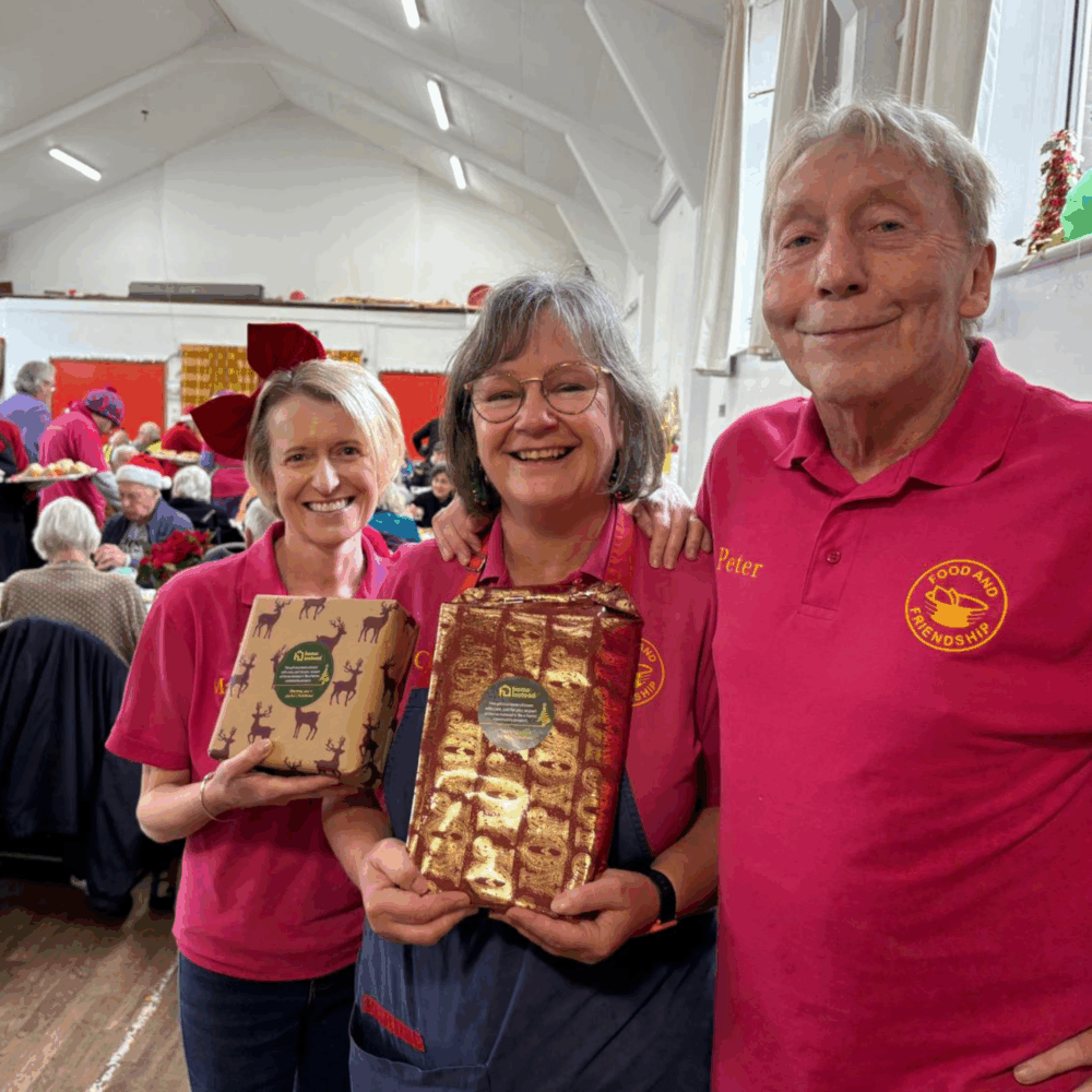 Three volunteers wearing pink “Food and Friendship” polo shirts smile at a festive indoor event, holding wrapped gift packages.