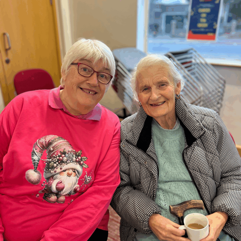Two smiling elderly women sitting together indoors, one wearing a pink Christmas sweater with a festive gnome design and glasses, the other in a mint green jumper and houndstooth jacket holding a white mug