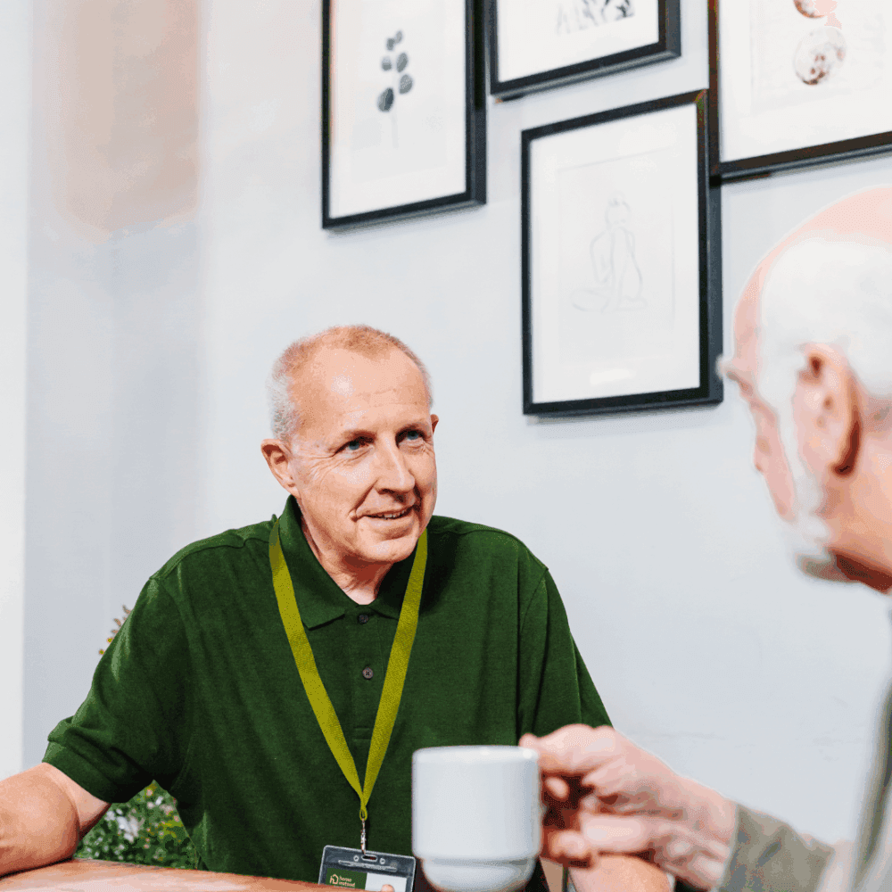 Two older men sit at a table, one holding a cup, talking in a room with framed artwork on the wall. - Home Instead