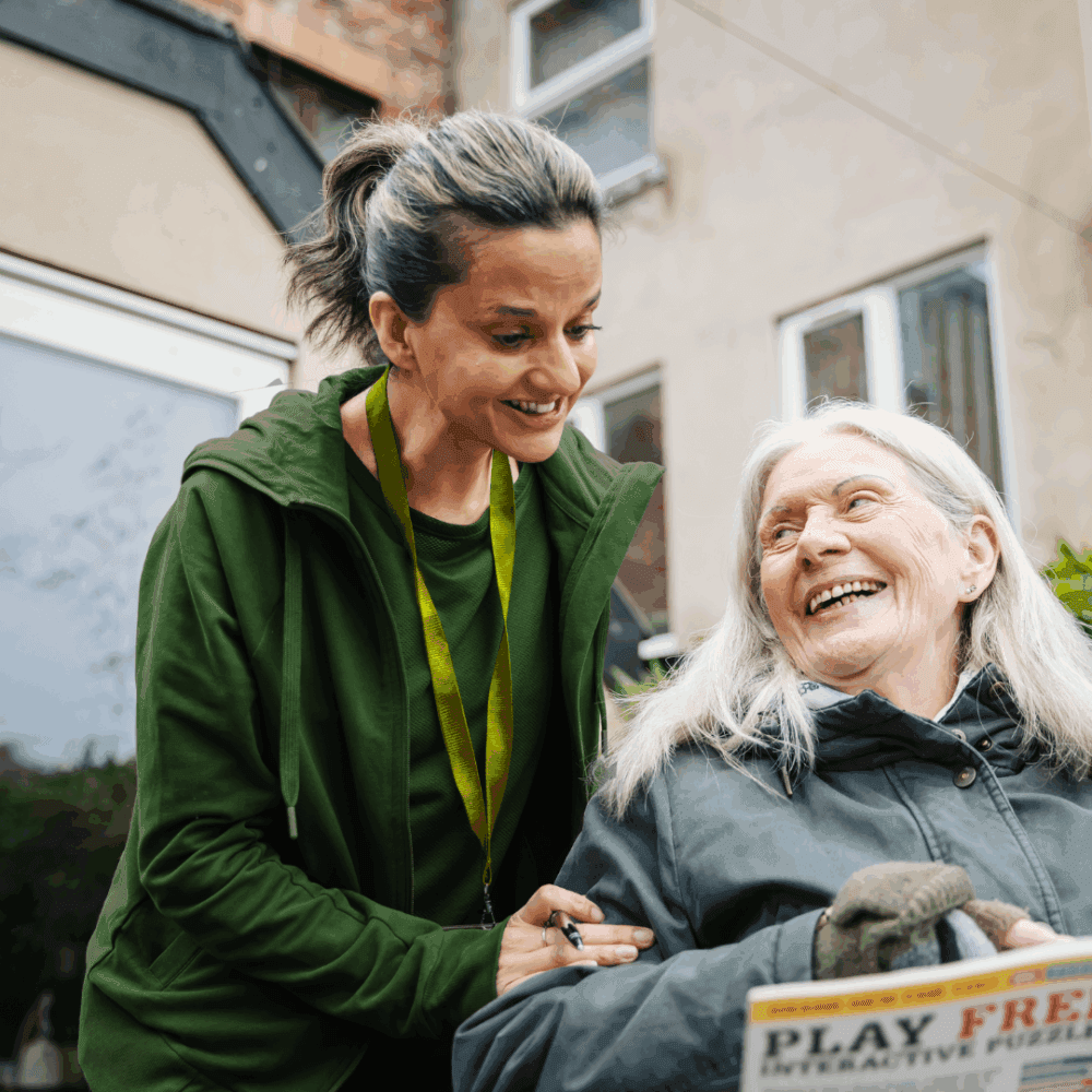 Young carer smiling at elderly woman in a wheelchair outdoors; both appear happy and engaged. - Home Instead