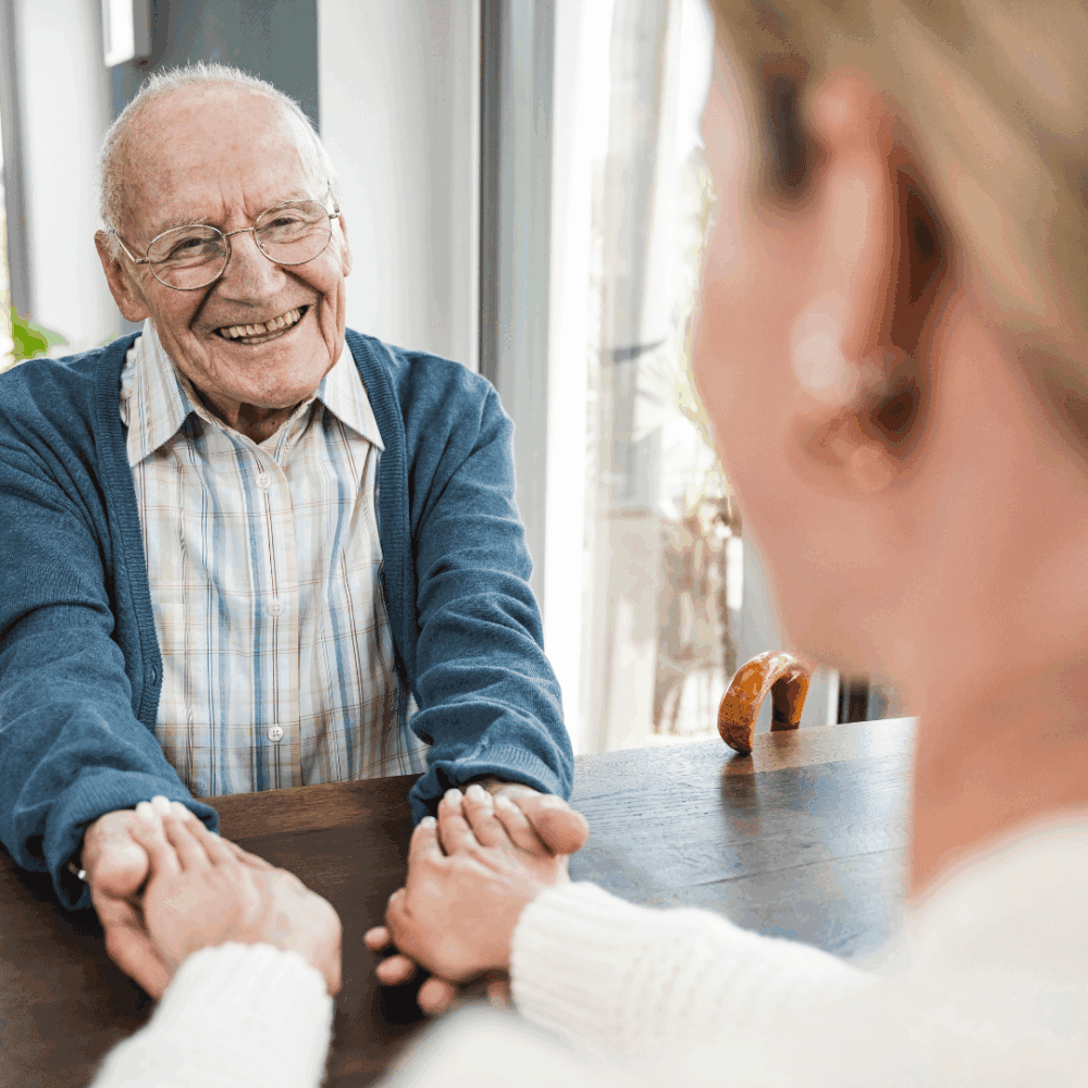 Smiling elderly man holding hands across a table with a younger woman in a bright, cosy room. - Home Instead