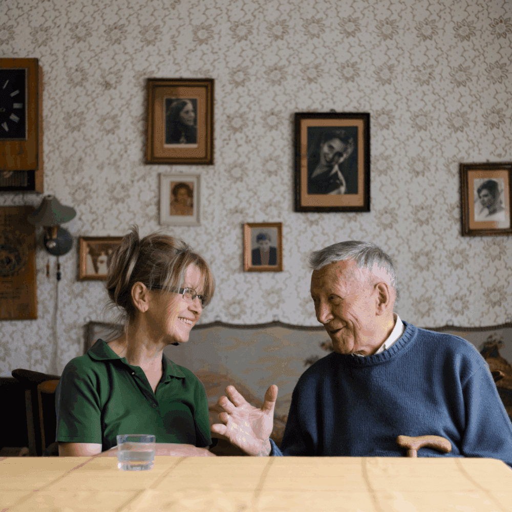 An elderly man and a woman smile and talk at a table in a cosy room with framed photos on the wall. - Home Instead