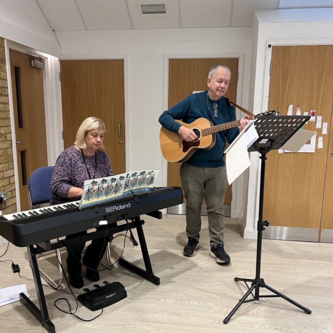 A woman plays a keyboard whilst a man stands next to her playing an acoustic guitar and singing. - Home Instead