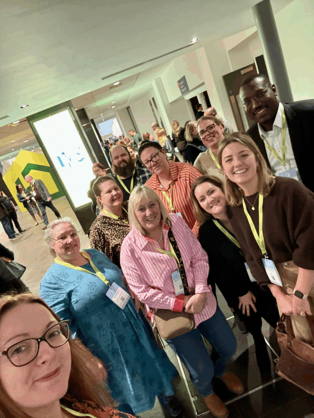 A group of people wearing lanyards smile for a selfie at an indoor event or conference. - Home Instead