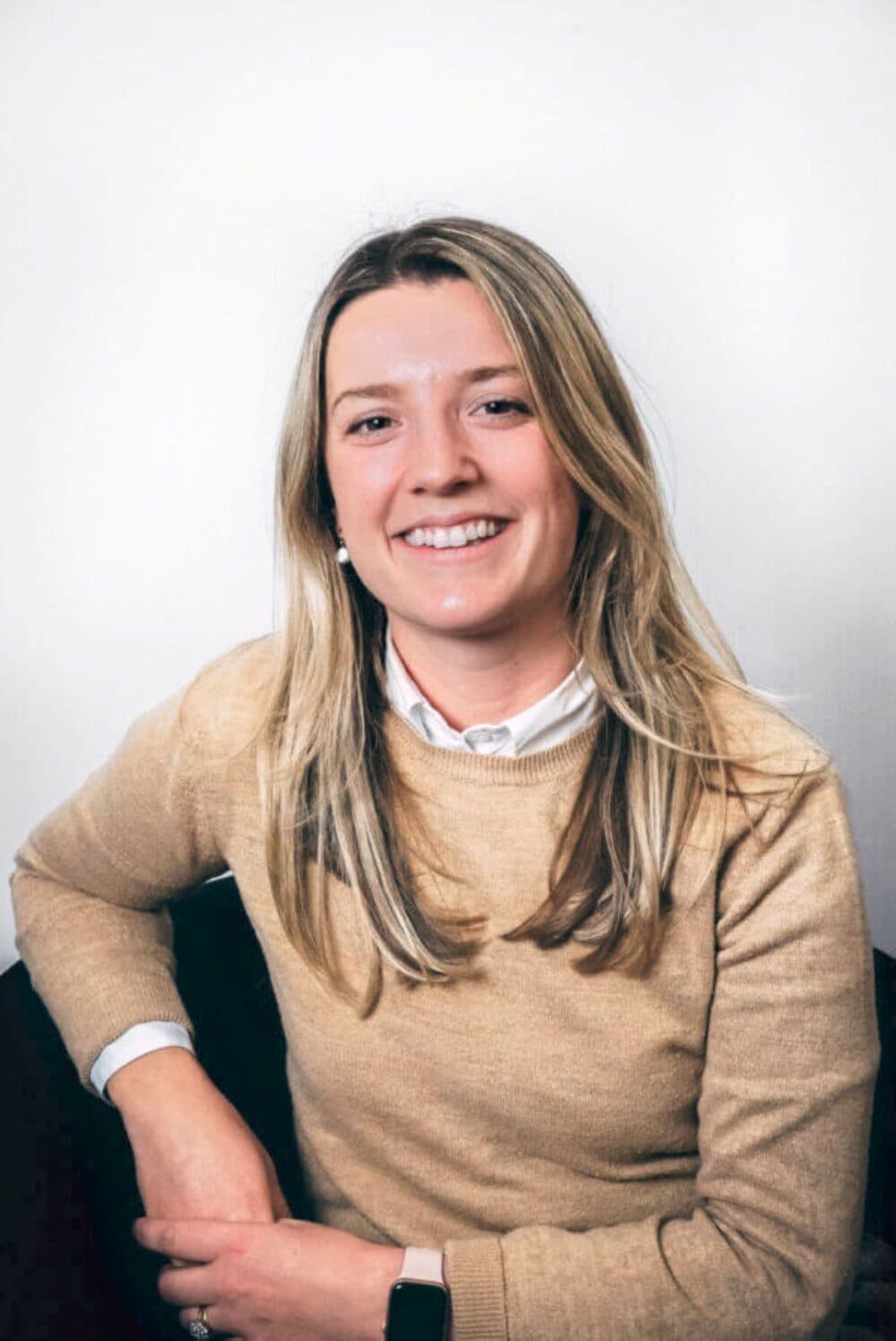 Woman with long blonde hair wearing a beige jumper and white shirt, smiling against a plain light background. - Home Instead