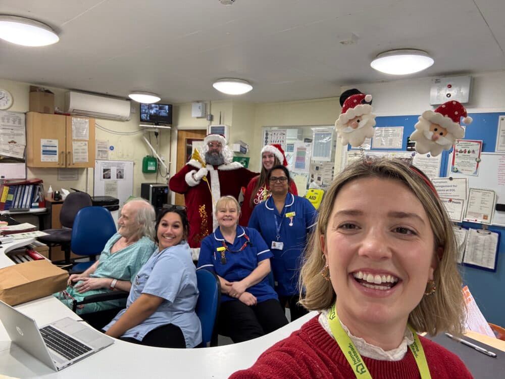 Six staff and a patient at a hospital desk; two people in Father Christmas costumes and one wears reindeer antlers. - Home Instead