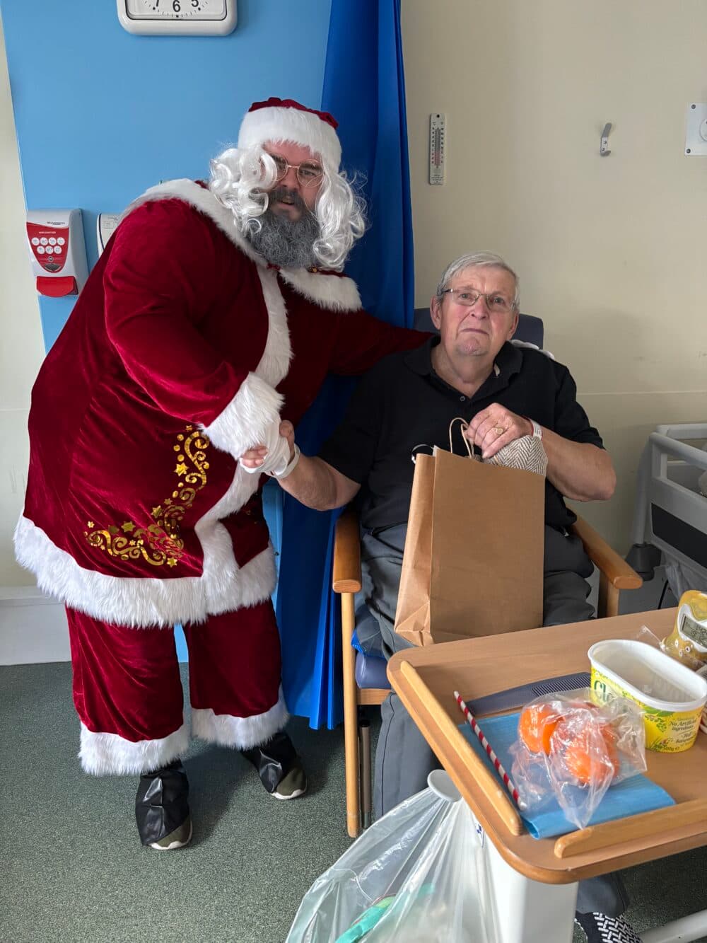 A man dressed as Father Christmas shakes hands with an elderly man in a hospital ward holding a gift bag. - Home Instead