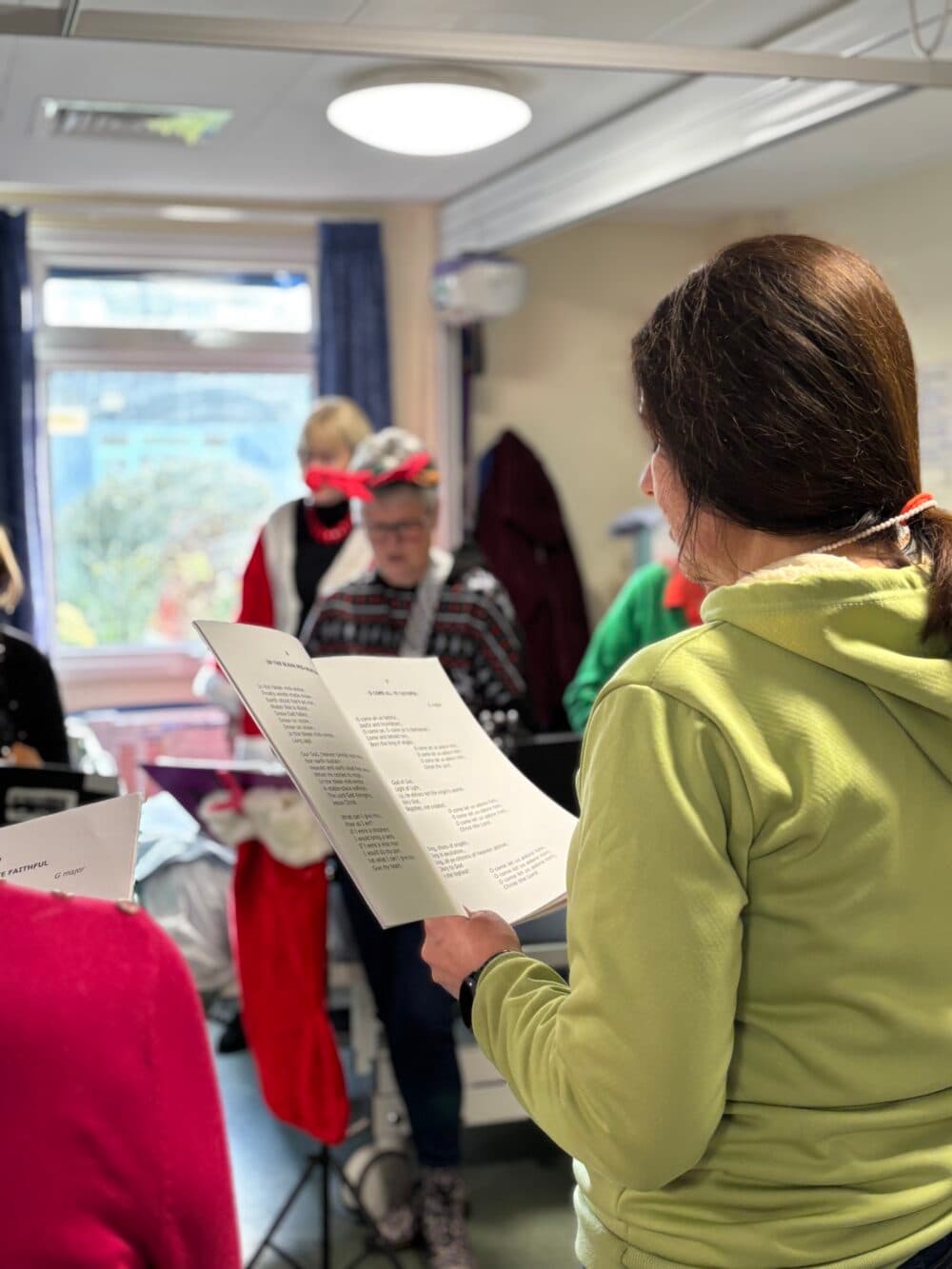 A group of people sing from song sheets in a brightly lit room, some wearing festive hats and jumpers. - Home Instead