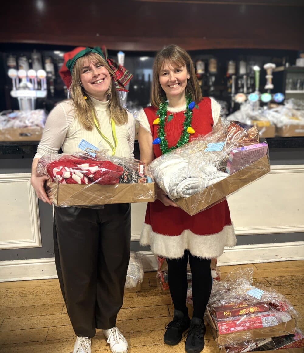 Two women in festive outfits smiling and holding gift hampers in a decorated indoor setting. - Home Instead