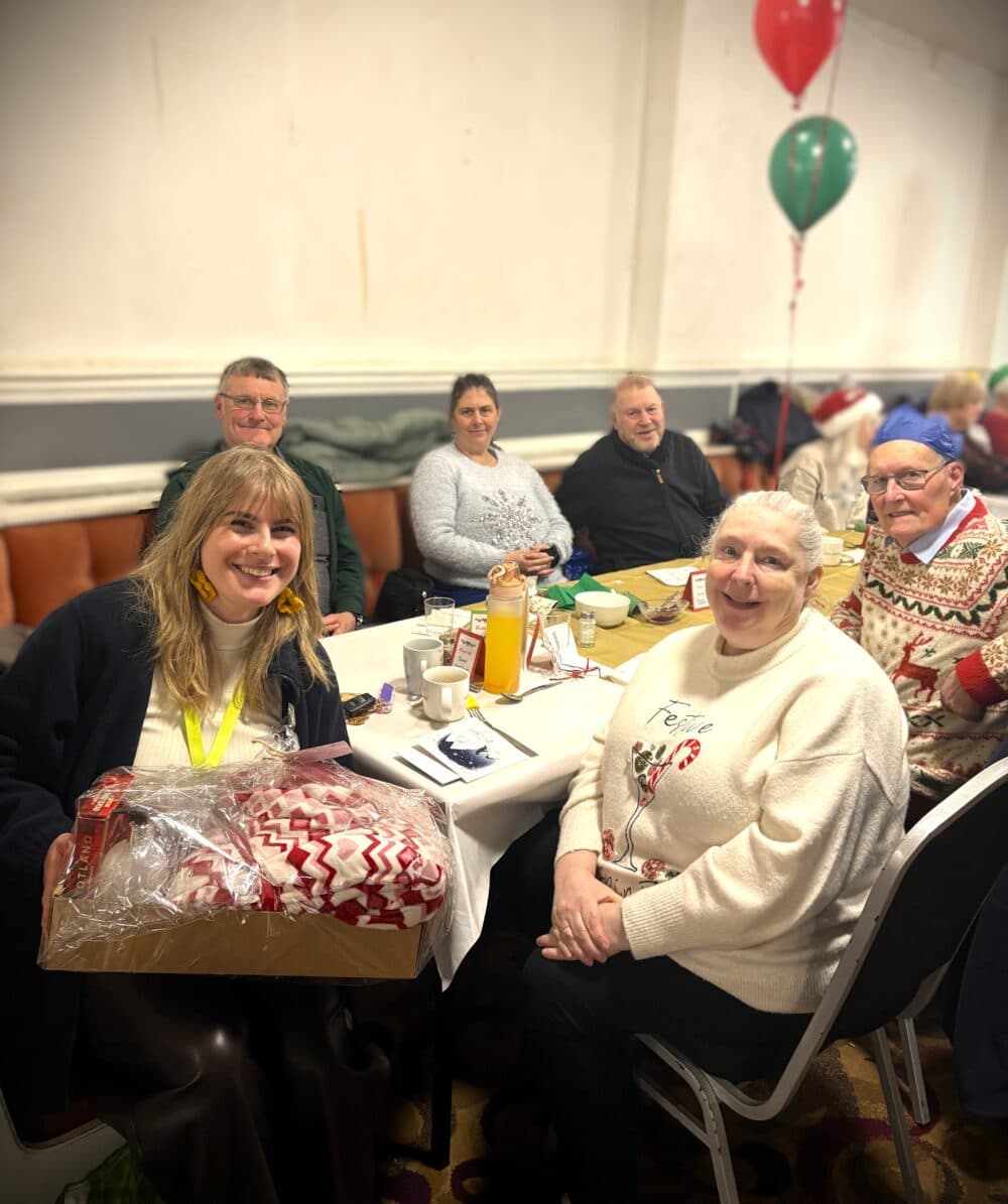 Six smiling people sit around a festive table, one woman holding a box with a red and white blanket. - Home Instead