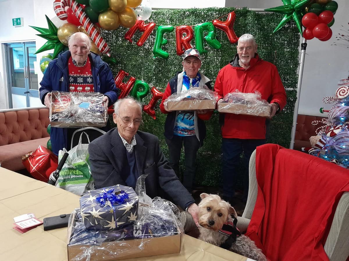 Four men with holiday gift hampers and a dog pose in front of festive decorations and a "Merry" sign. - Home Instead