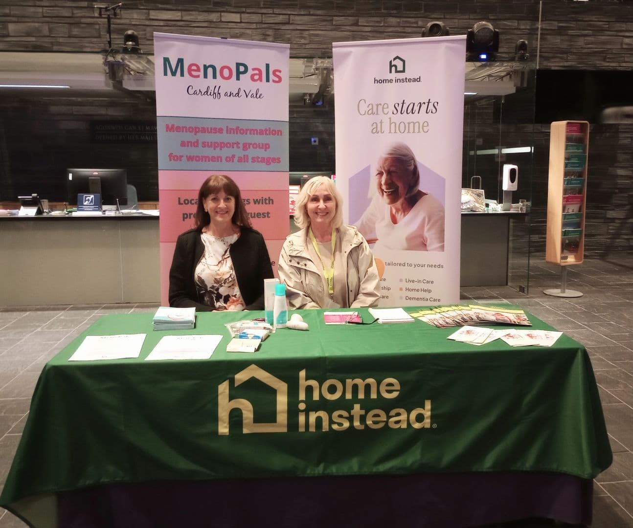Two women sit at a table with Home Instead and Menopals banners, offering menopause and home care information. - Home Instead