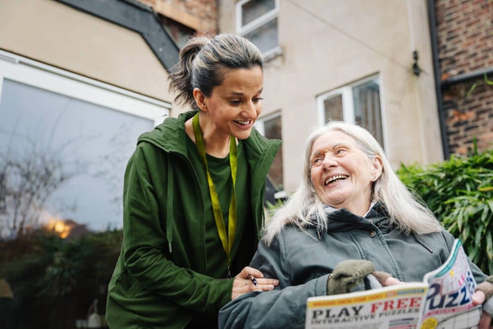 A younger woman smiles at an older woman in a garden as the older woman holds a puzzle magazine. - Home Instead