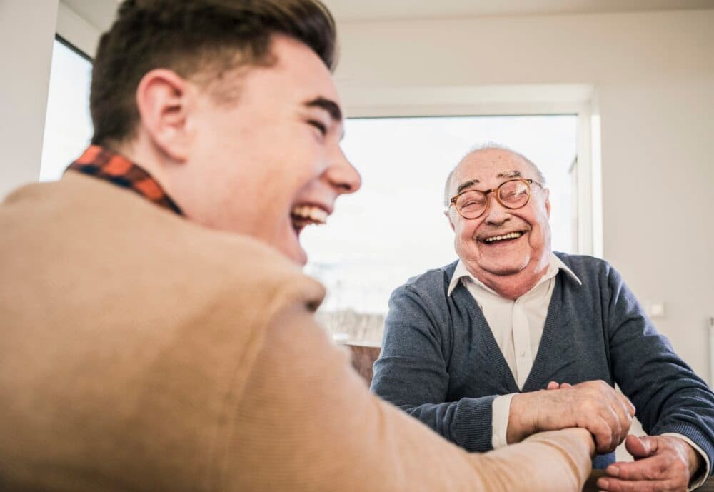 Young man and elderly man laughing together at a table in a bright room with a window. - Home Instead