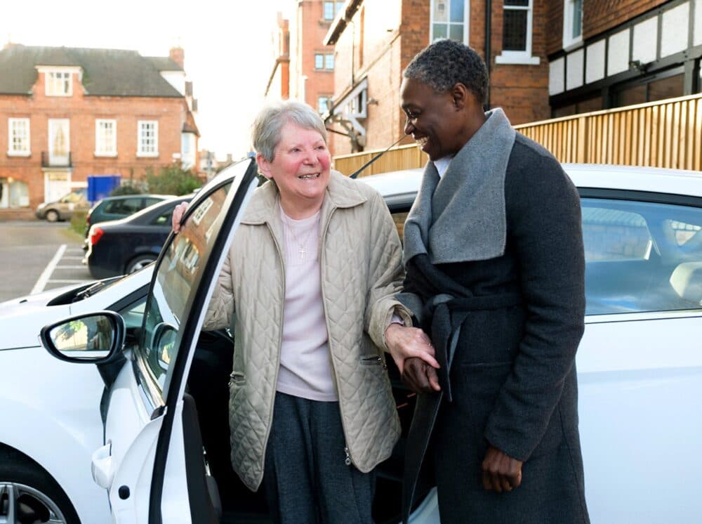 Two women smile at each other as one helps the other out of a parked white car in a residential area. - Home Instead