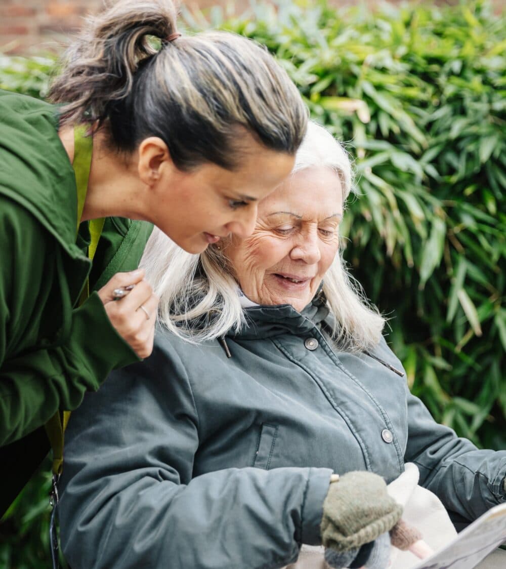 A younger woman and an older woman smile whilst looking at a newspaper together outdoors. - Home Instead