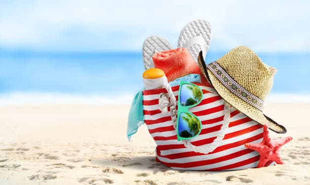 Striped beach bag on sand with sunglasses, hat, flip-flops, towel, suncream, and a starfish by the sea. - Home Instead
