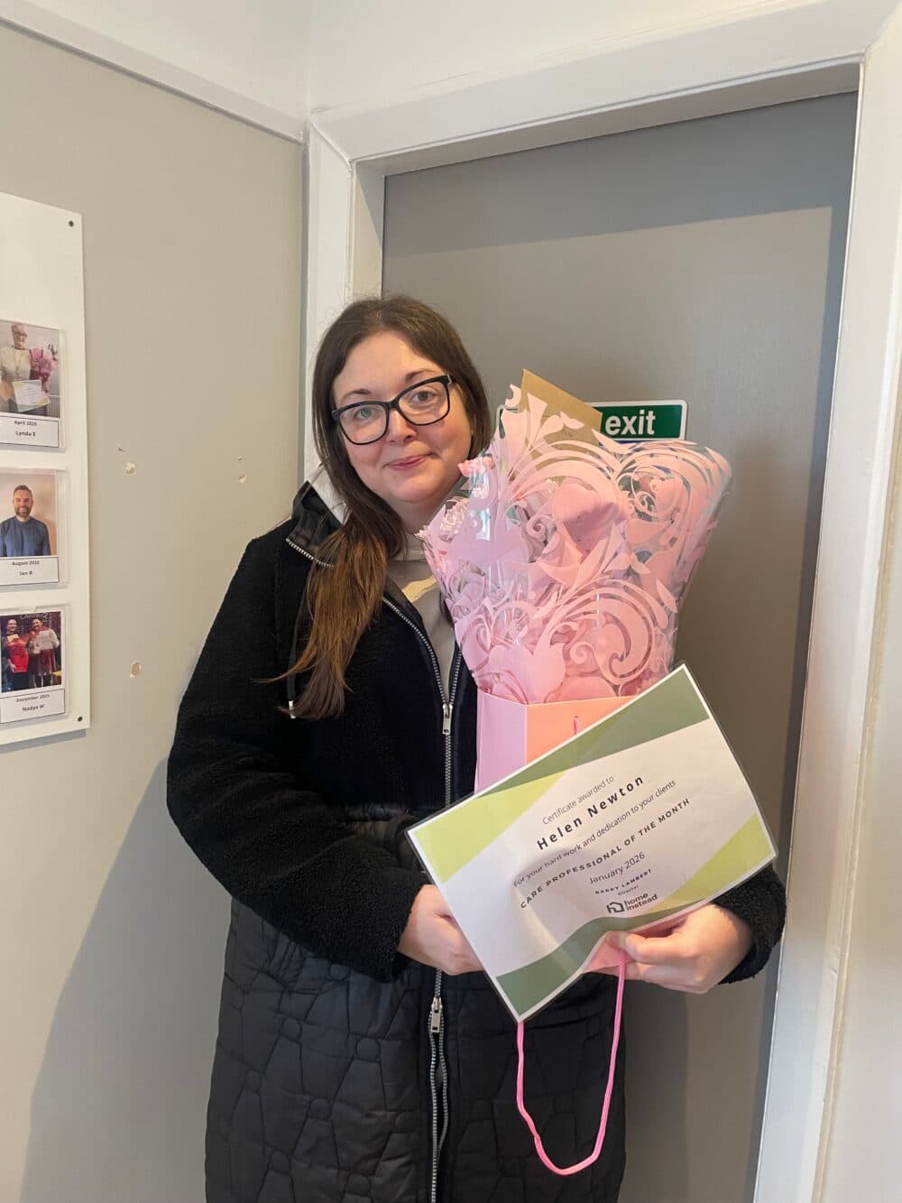 Woman holding a bouquet and a certificate, smiling whilst standing indoors near a door and wall display. - Home Instead
