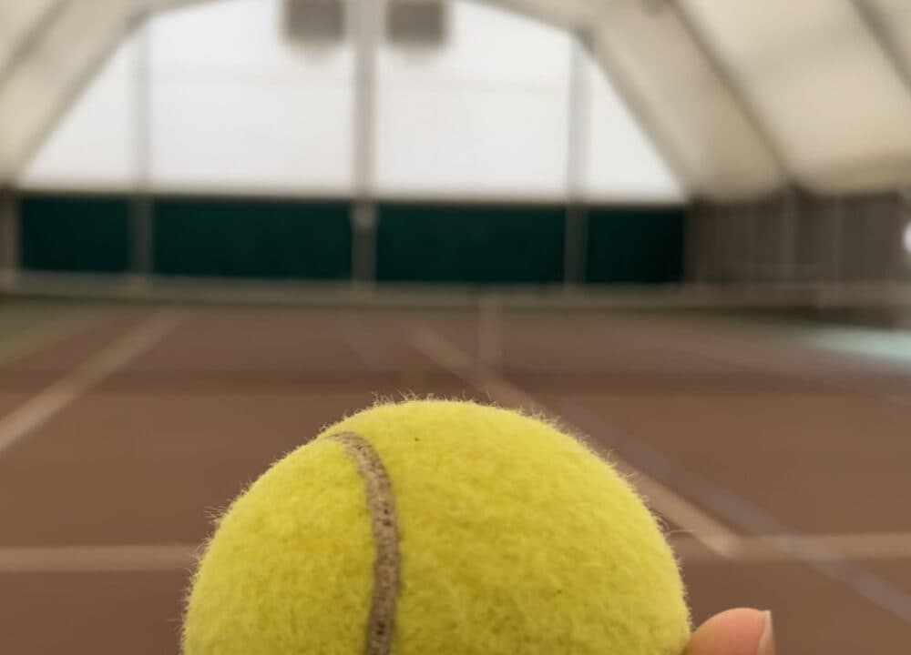 Close up of a tennis ball being held up against the background of an indoor tennis court.