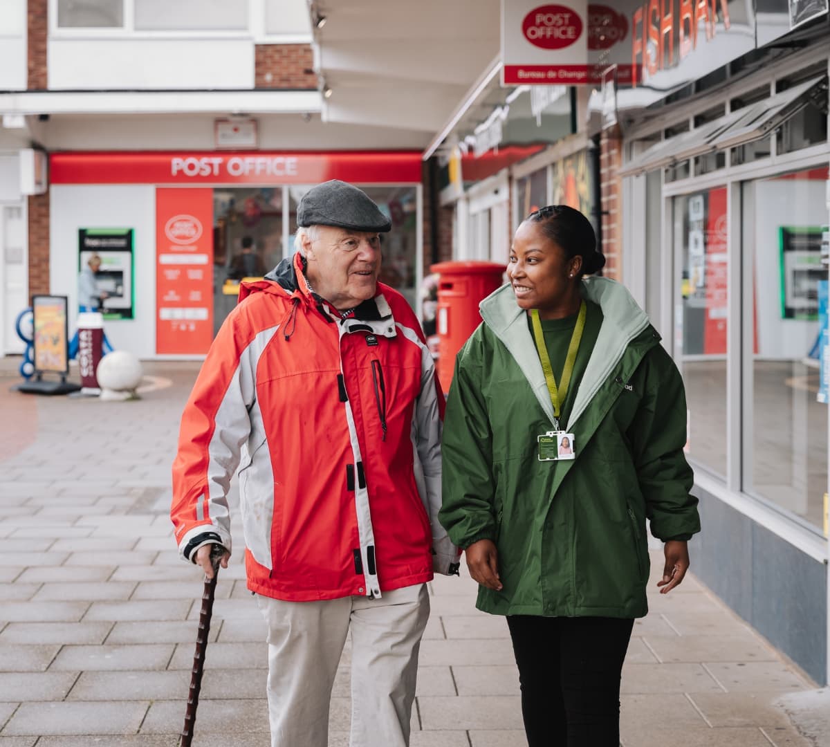 An older male adult wearing jacket and a hat walking using a crane with his younger female carer wearing green jacket