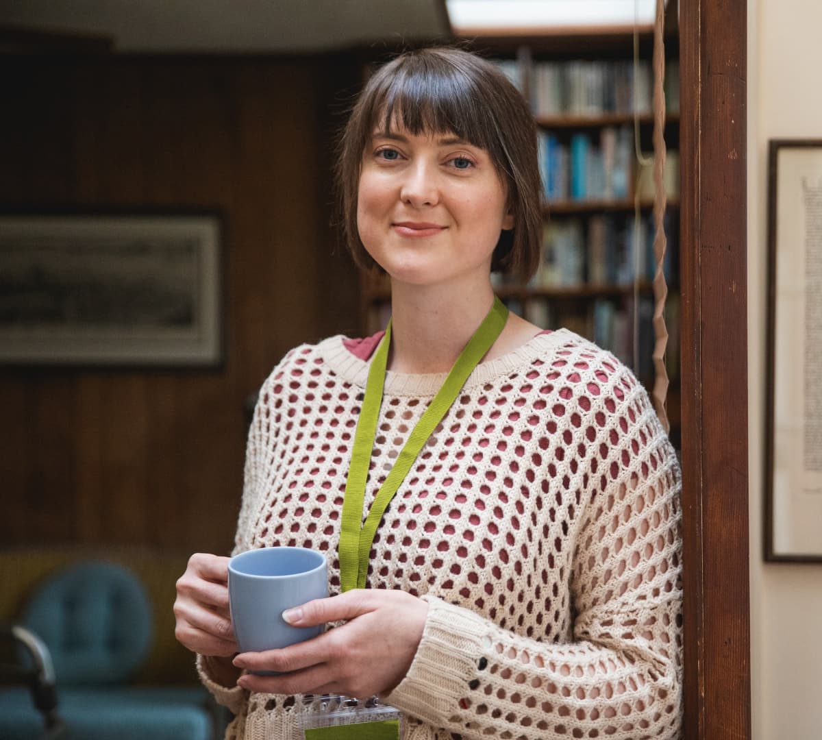 A woman with short hair smiling and standing while holding a cup of coffee inside the house