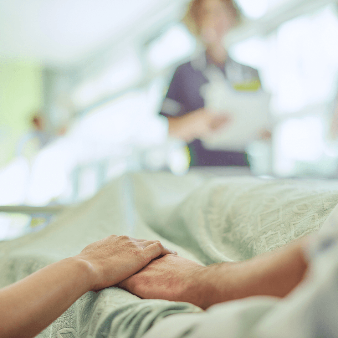 A close-up, poignant shot of two hands clasped together over a hospital bed cover, symbolizing support and advocacy, with a blurred healthcare professional working in the background.