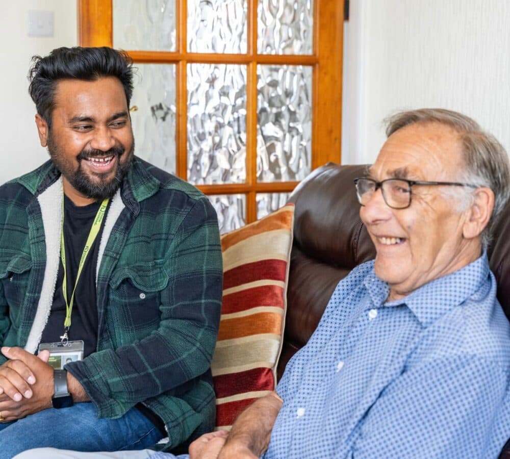 An older male adult with grey hair and wearing eyeglasses happy and smiling with his younger male carer with black hair both sitting on the couch