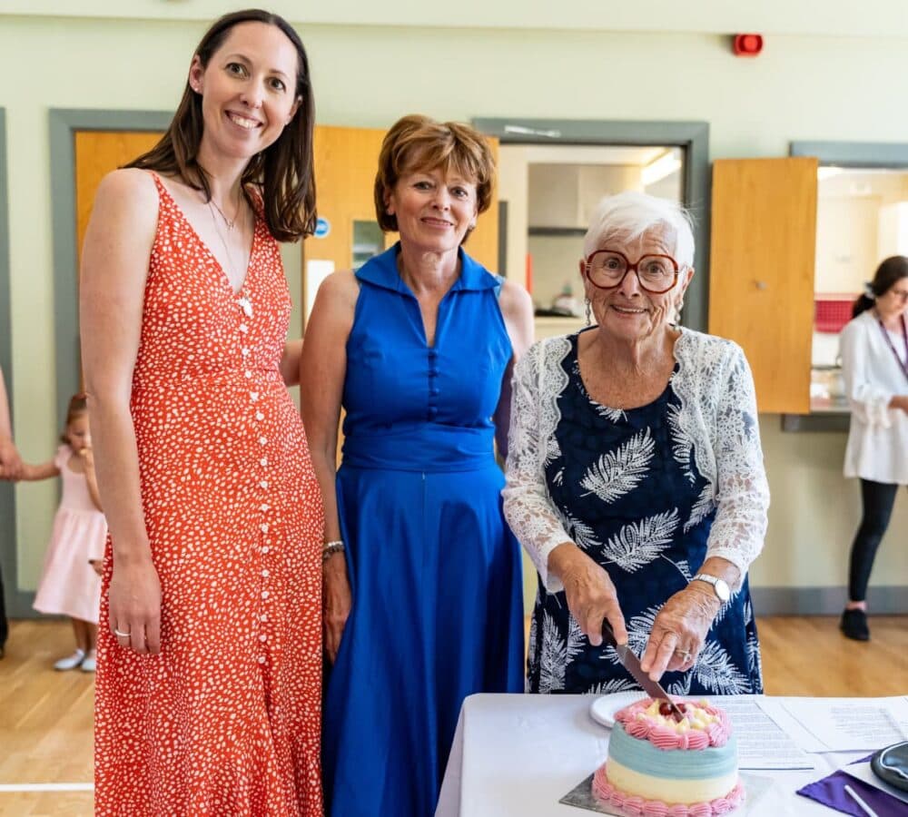Three women standing and smiling while the oldest one is slicing a cake on the table - Home Instead