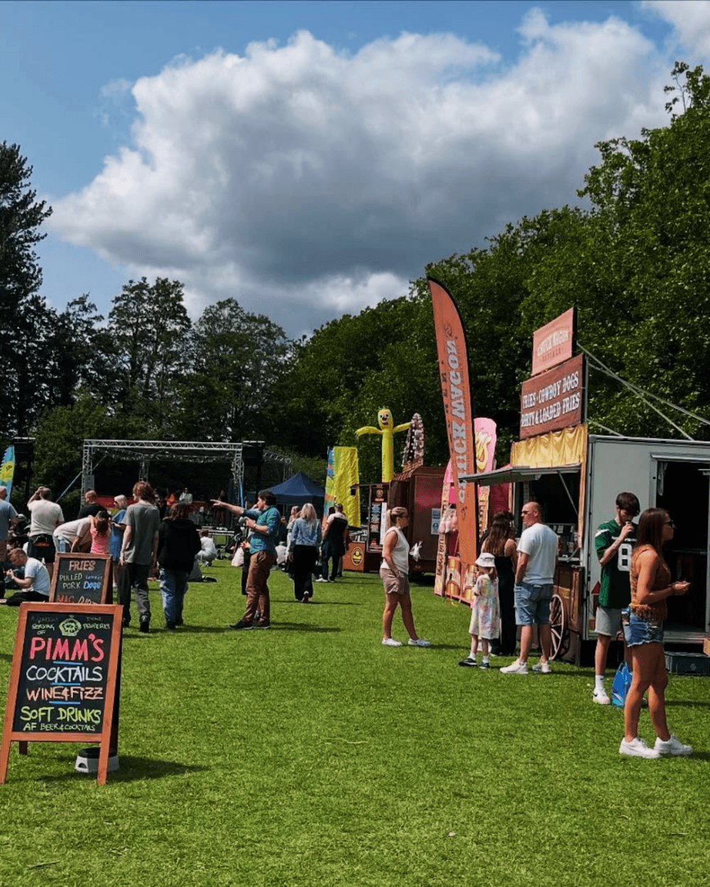 People at an outdoor festival with food stalls, signs, and trees under a partly cloudy sky. - Home Instead