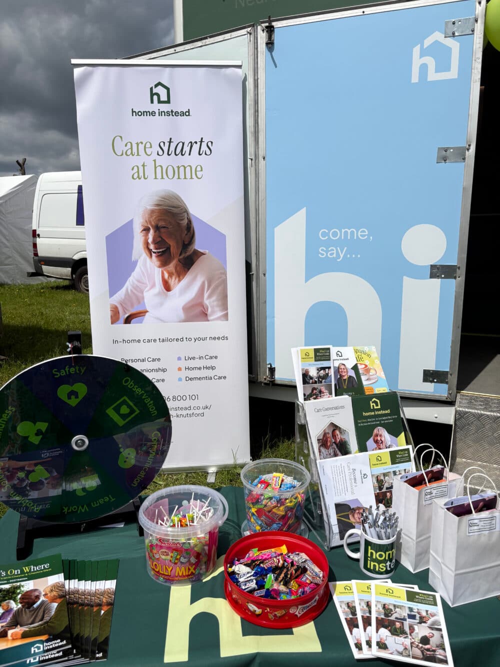 A table with leaflets, sweets, and a prize wheel at a "Home Instead" care service event stall outdoors. - Home Instead
