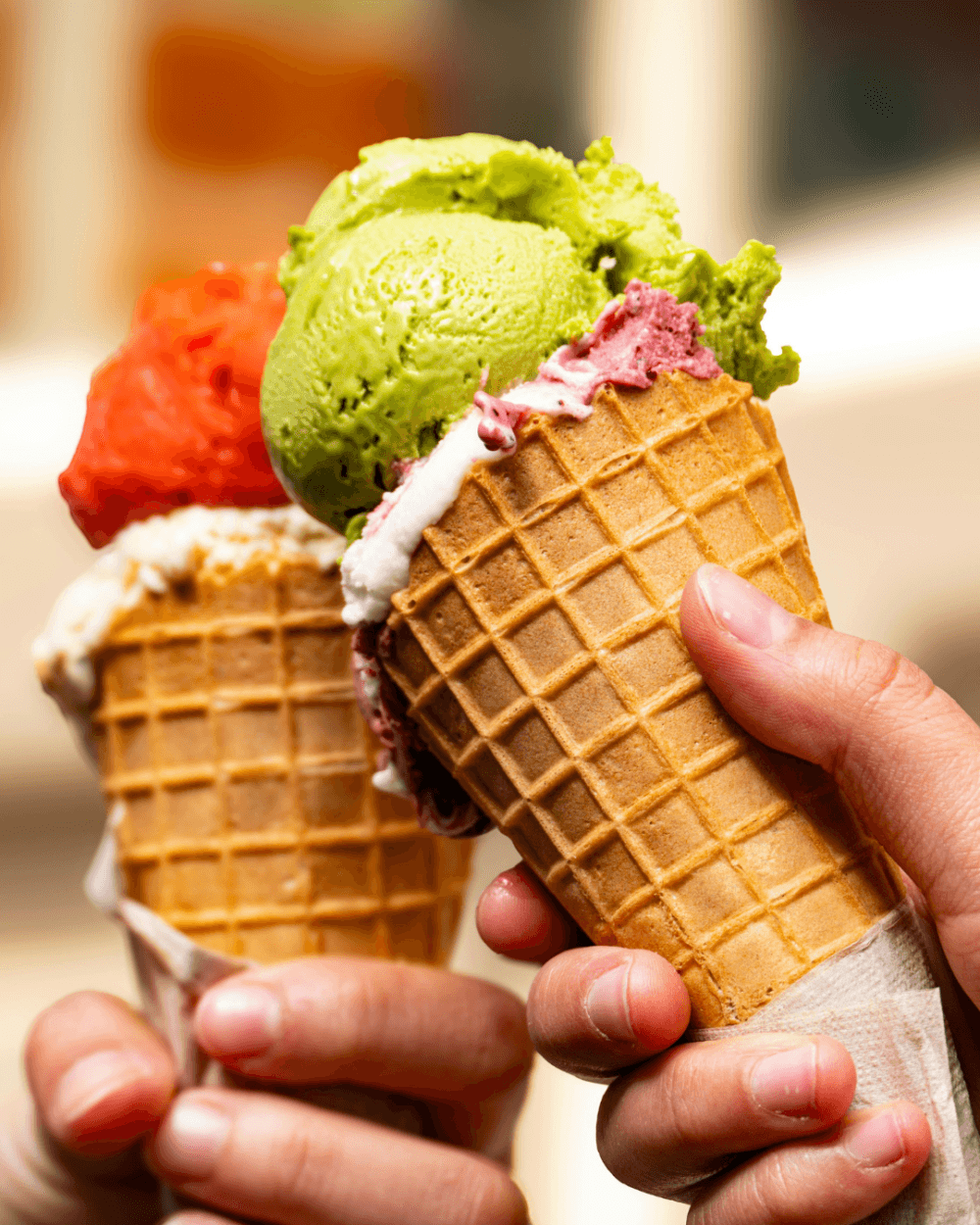 Two hands holding waffle cones with colourful scoops of ice cream in green, red, pink, and white flavours. - Home Instead