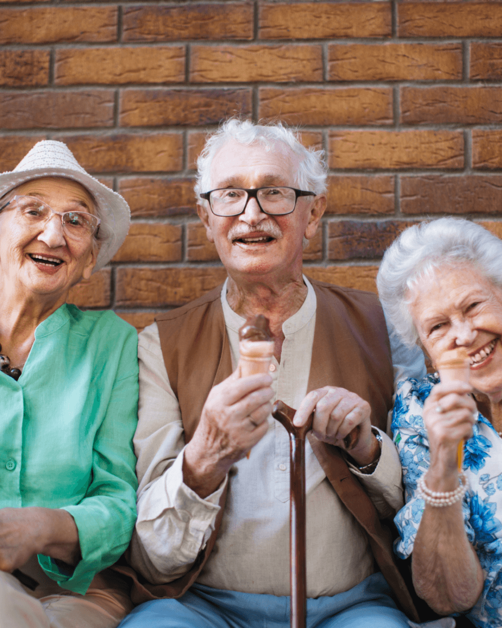 Three smiling elderly people sit against a brick wall, enjoying ice cream cones together. - Home Instead