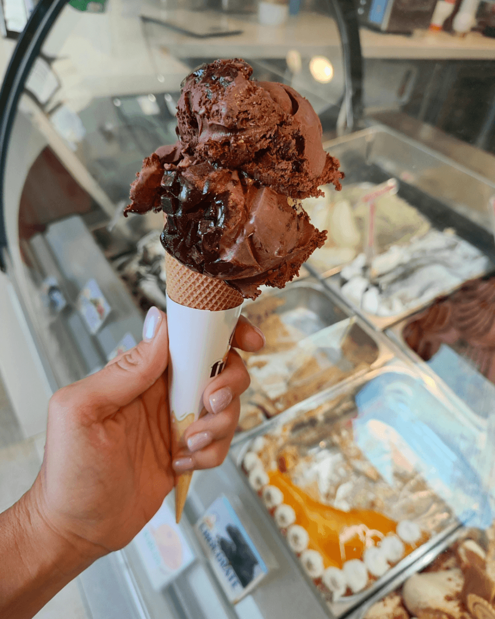 A hand holds a chocolate ice cream cone inside a gelato shop with tubs of gelato in the background. - Home Instead